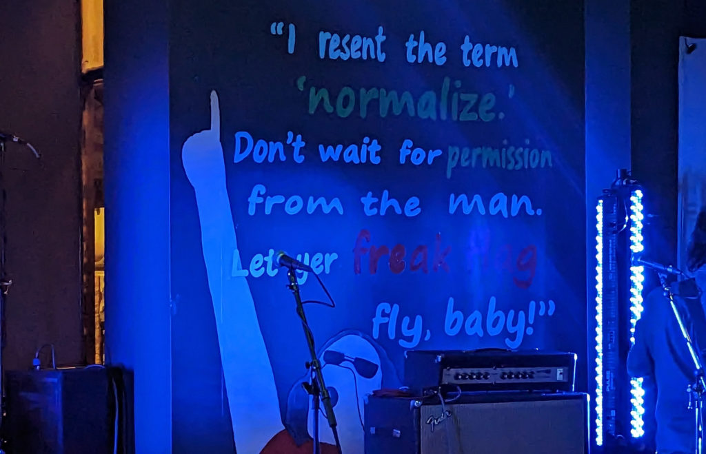Photo of a mural behind the stage at The Space. It features a painting of a singer pointing to the sky with a mic to his mouth, and above him the quote: "I resent the term 'normalize'. Don't wait for permission from the man. Let yer freak flag fly, baby!"