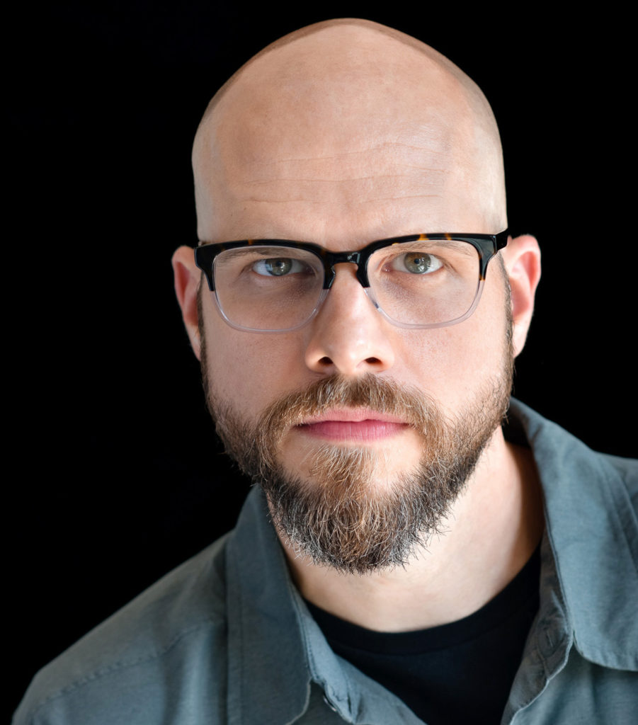 A headshot of Vincent Carlson. He is a white man, bald, facial hair, wearing glasses. He is looking straight at the camera with a serious expression.