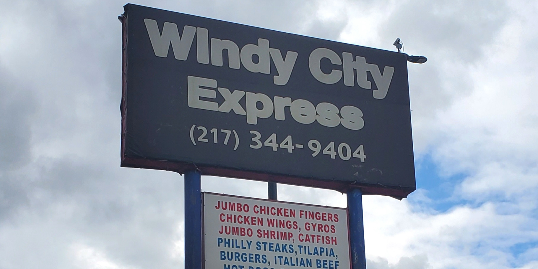 Windy City Express in Urbana's sign on a cloudy blue day. The sign also has the fast food restaurant's phone number and below it a list of the popular dishes. Photo by Carl Busch.