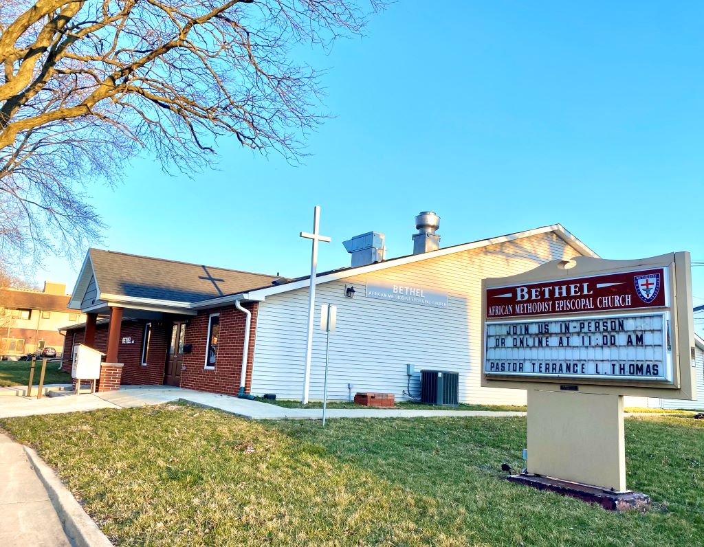 A one story building with red brick on one side, and light beige siding on the other. There is a sign in the grass in the foreground that says Bethel African Methodist Episcopal Church.