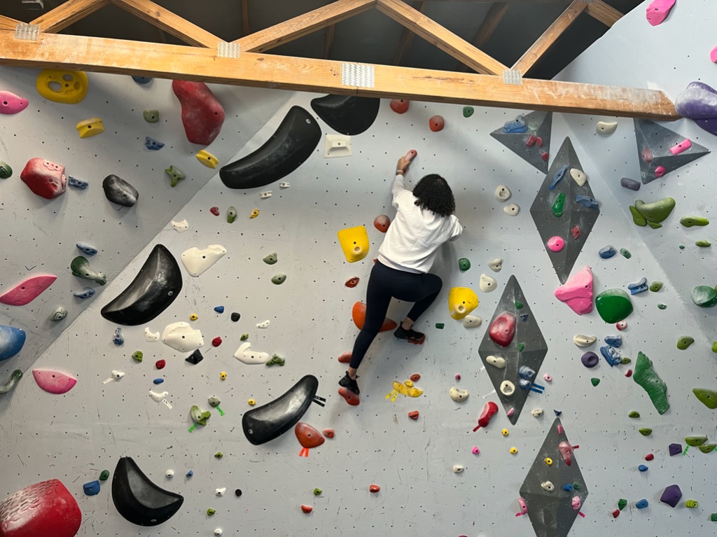 a tan woman with dark curly hair climbs up a grey rock wall with colorful rocks. she is wearing a white sweatshirt, navy leggings, and rock climbing shoes.