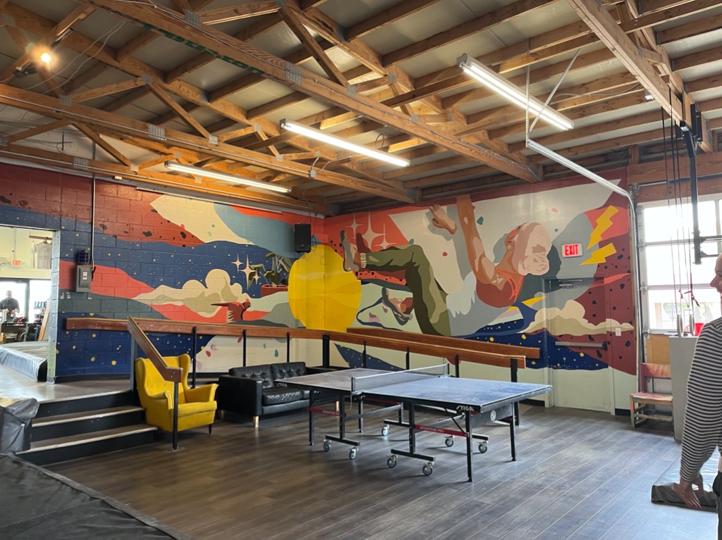 a big room with a ping pong table in the foreground a yellow chair, and a black couch. The wall has a brightly colored mural with a female rock climber.