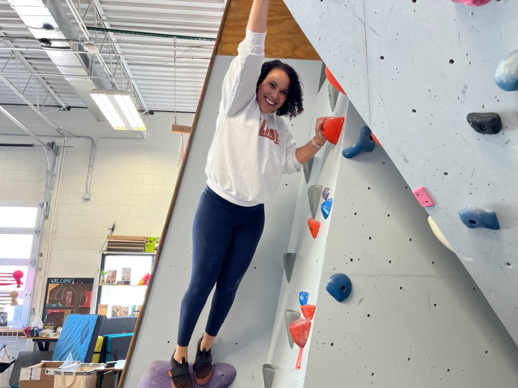 a tan woman with dark curly hair is stretched out reaching for a climbing rock. she is wearing a white sweatshirt with the words Illinois, navy leggings, and rock climbing shoes. she is laughing.