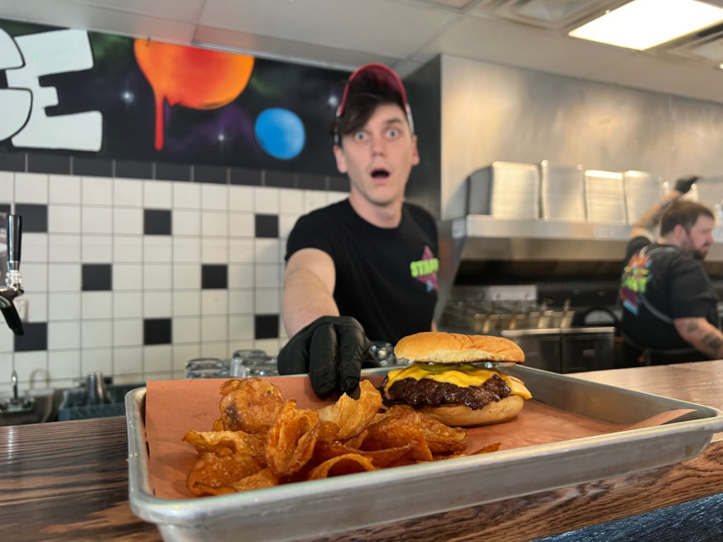 A person who appears to be a chef presenting a tray with a burger and fries to the camera