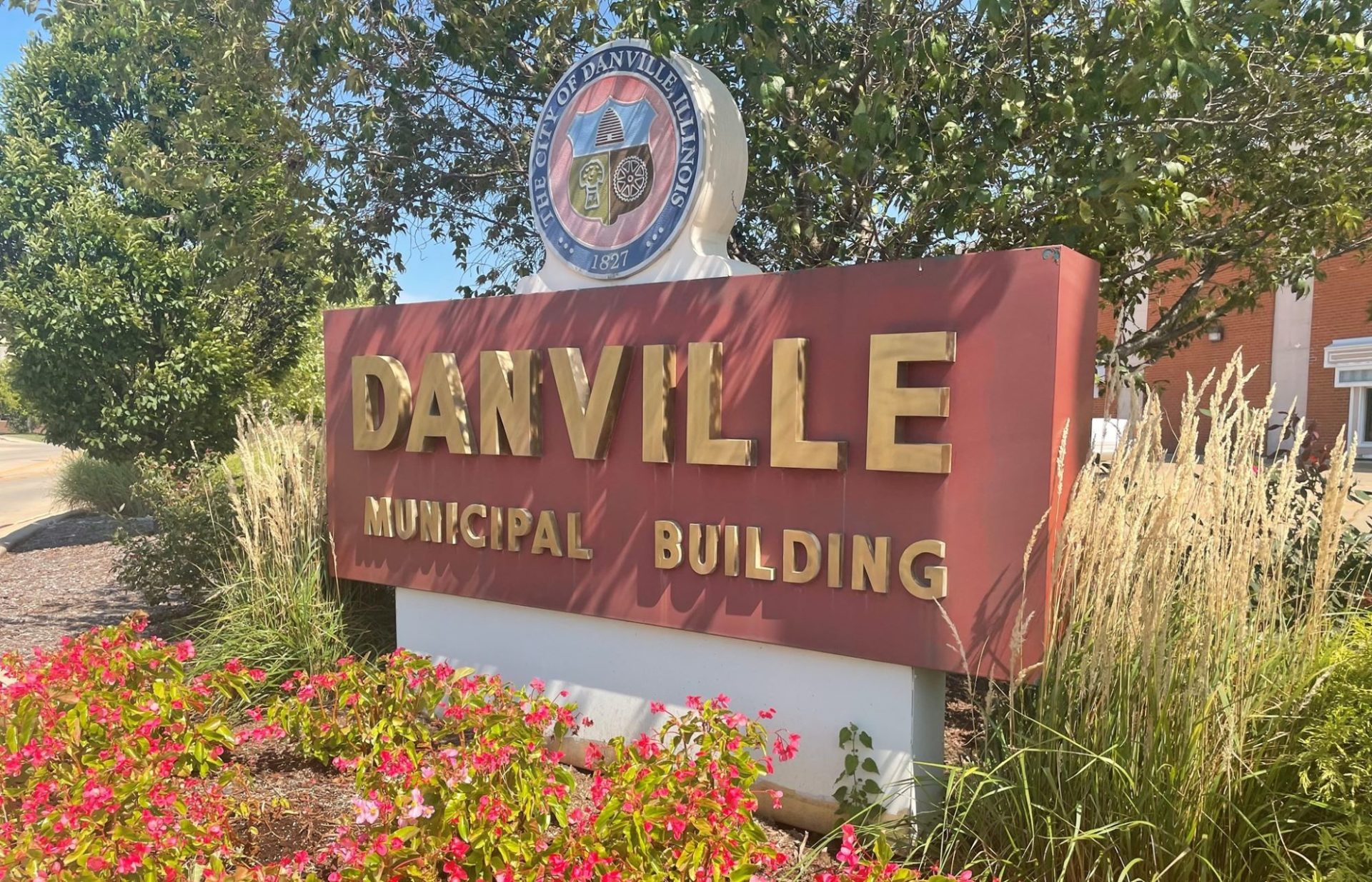 A brick red rectangular sign that says DANVILLE MUNICIPAL BUILDING in gold letters. It is surrounded by flower beds and decorative grasses.