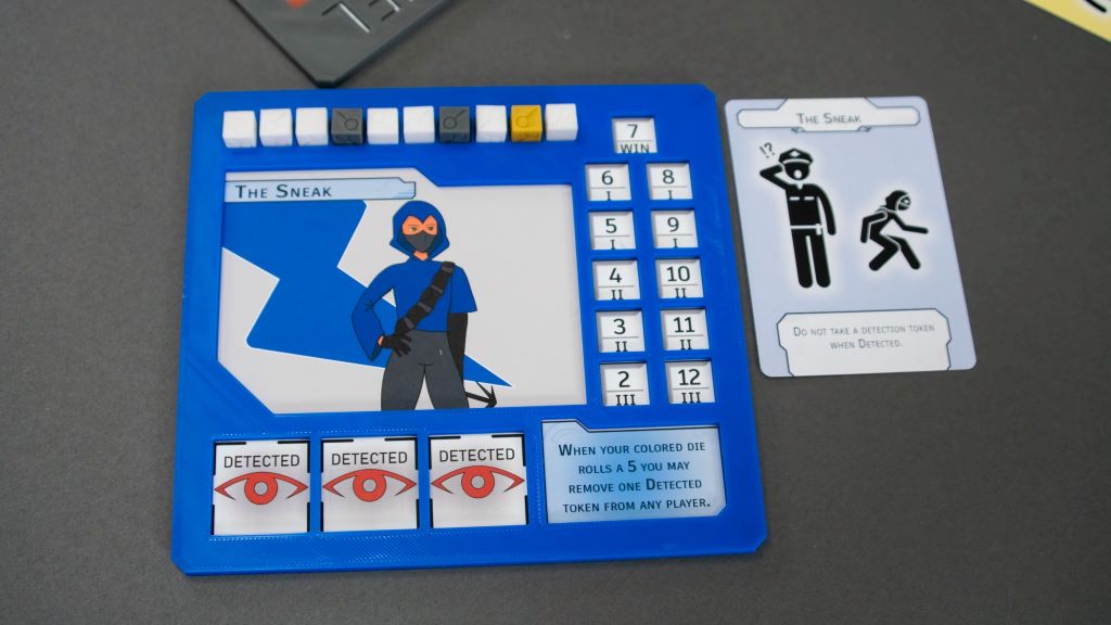 A square game that is blue with white, black, and yellow square buttons along the top and an image of a ninja below. Along the bottom are three red eye images with the word "detected" over them. There is a card with two black stick figures sitting next to the game.