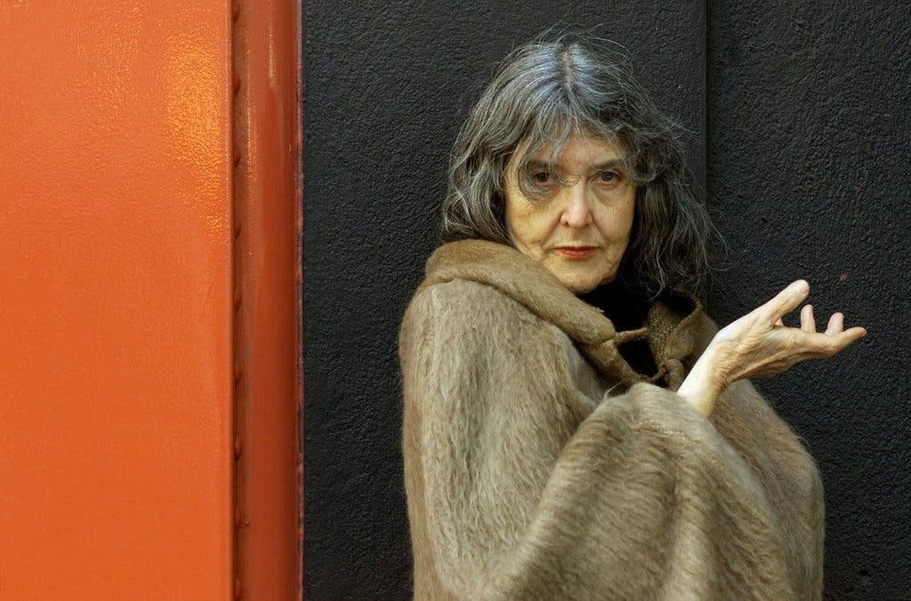 A photograph of María Irene Fornés. A Cuban woman wearing a fur coat, with one hand held int he air. She has long grey hair, and is leaning against a bright orange wall.