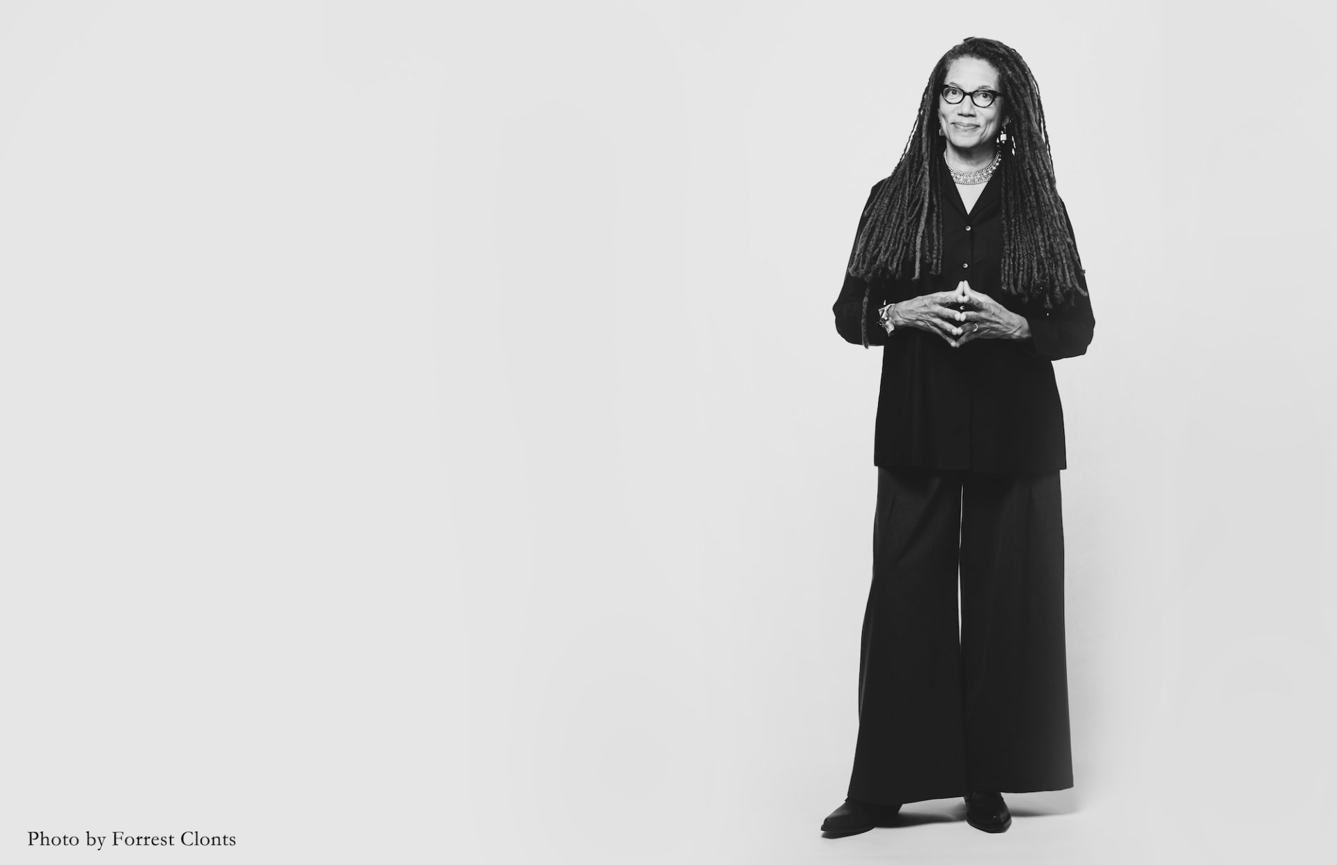Nikky Finney stands on the right side against a plain white background. She wears black wide -legged pants and a long sleeve black top. Her arms are bent with hands touching in the middle. She has dreadlocks extending to her waist.