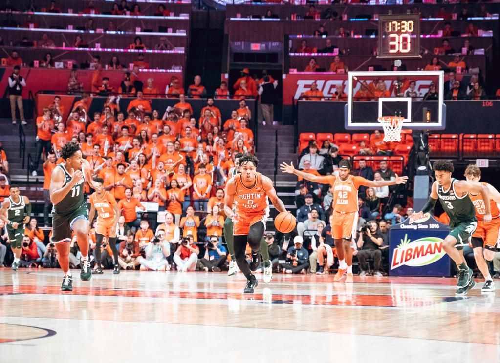 A black man in an orange jersey dribbles a basketball down the court towards the camera. Other players and fans watch from a distance.