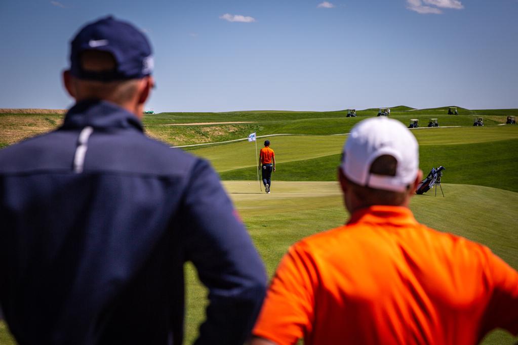 Two men stare at a golfer in the distance walking up to the pin. The man on the green wears a red shirt while the men in the foreground wear an orange polo and white hat and a blue long sleeve shirt with a blue hat.