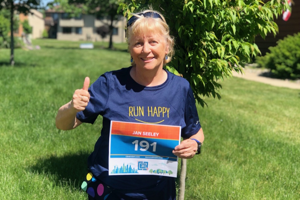 a woman with blonde hair in a ponytail is wearing a blue shirt that says run happy. She is holding a runners bib. She is smiling and giving a thumbs up to the camera. 