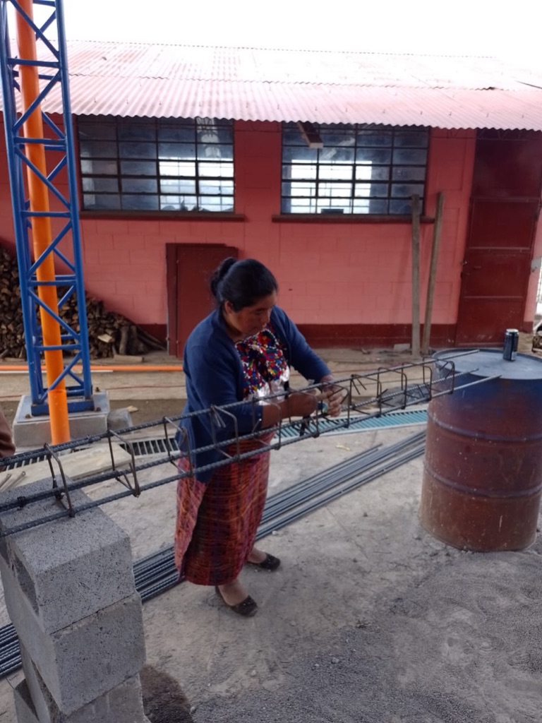 a woman in a blue sweater, floral print shirt, and long red skirt stands in front of a red building with a tin roof. She is connecting rebar which is laid across cinder blocks and an oil drum.