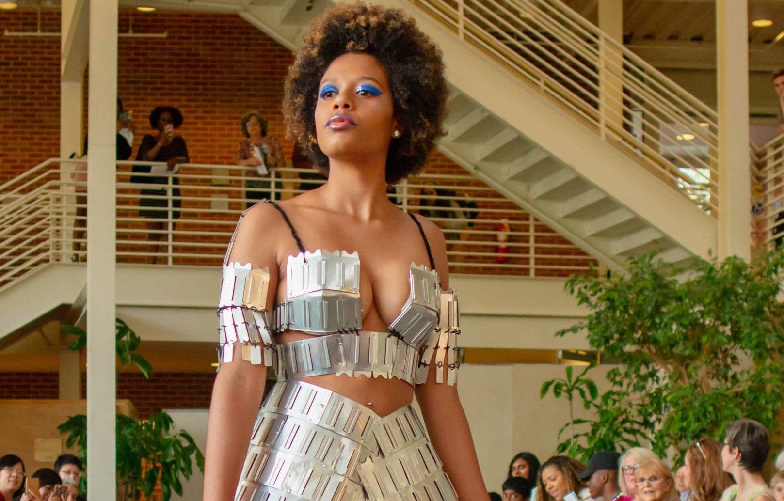 A runway model. A Black woman models a silver metallic top and skirt. The top is off the shoulder. The skirt tapers in the middle. She is wearing blue eyeshadow.