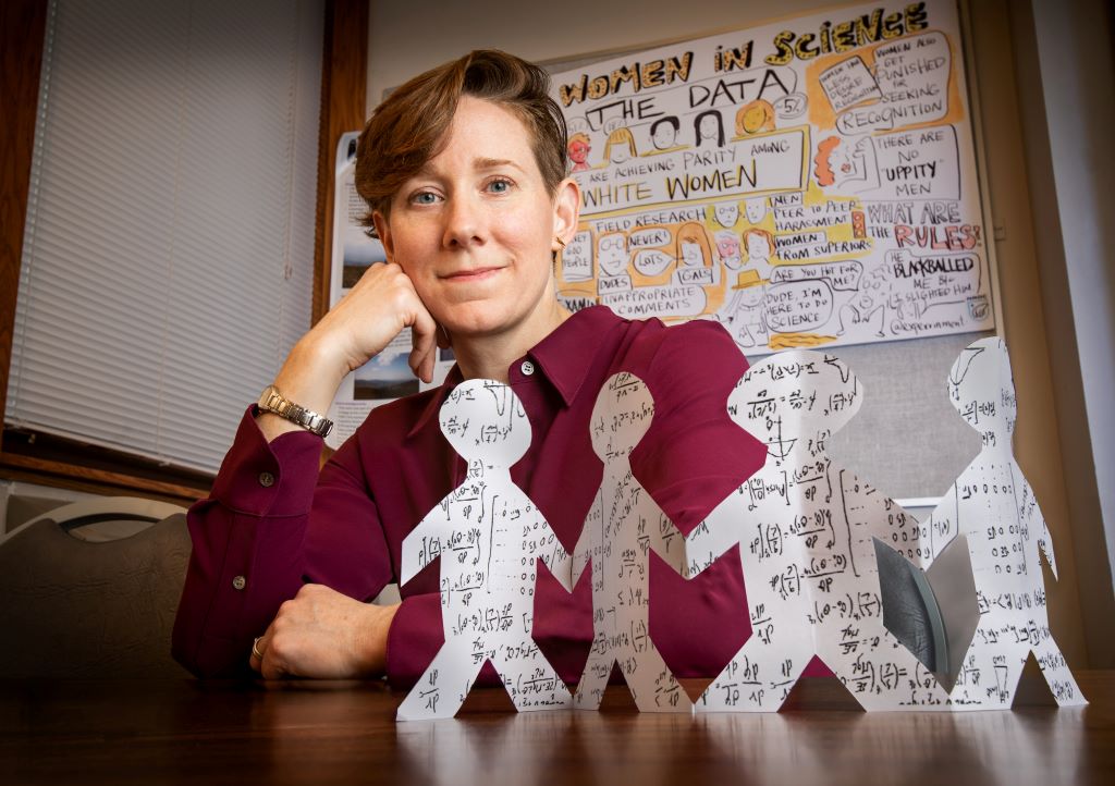 A woman with short brown hair is wearing a dark red collared shirt. She is sitting at a table, with her hand resting on her chin. There are paper doll cutouts in front of her.