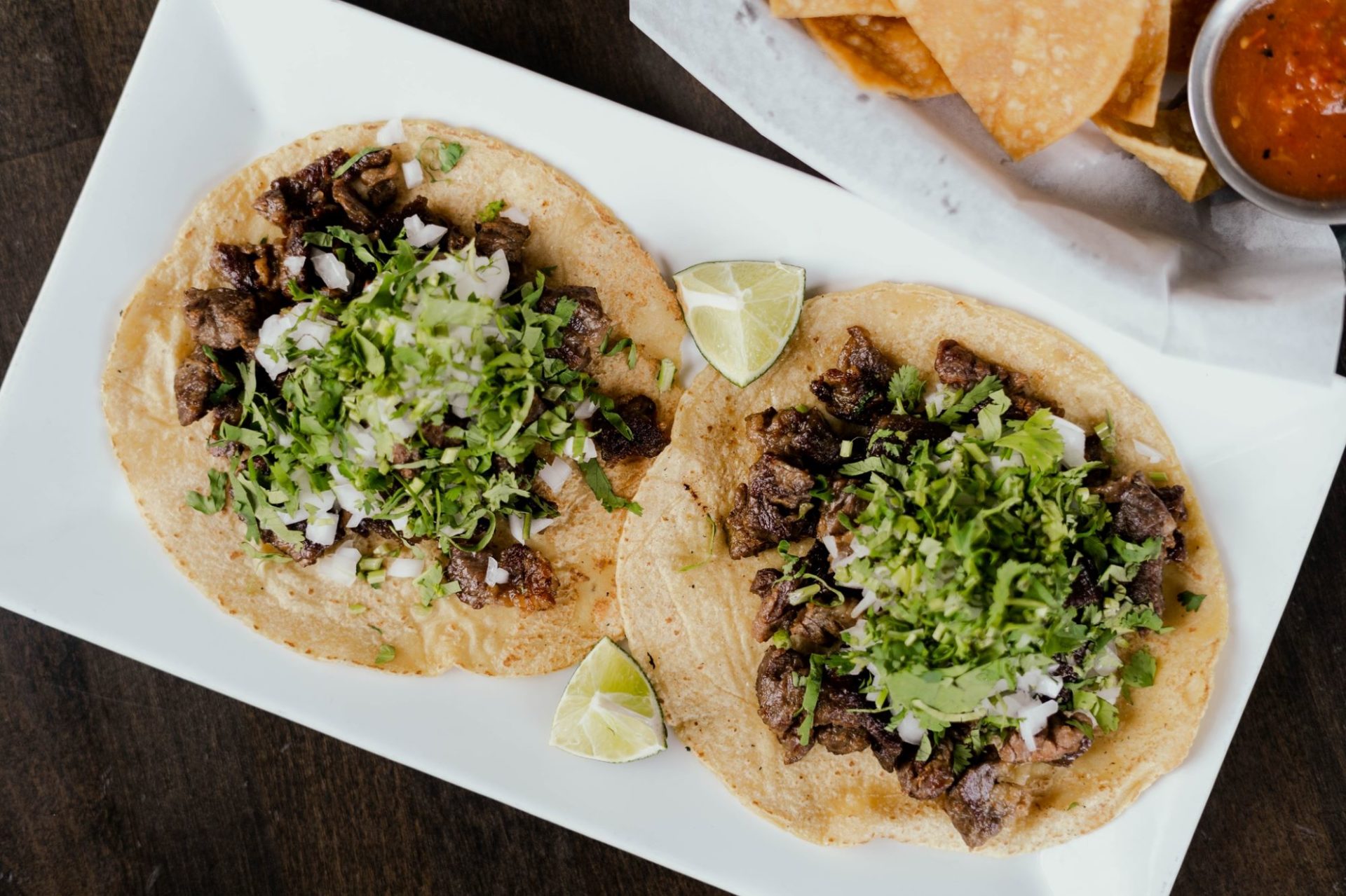 Two tacos on round tortillas with steak, onion pieces, and green cilantro sit side by side on a white rectangular plate.