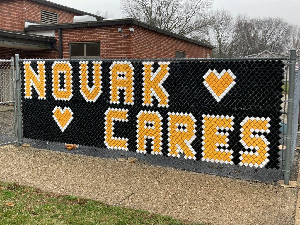 A fence with a large black sign with orange letters that say NOVAK CARES.