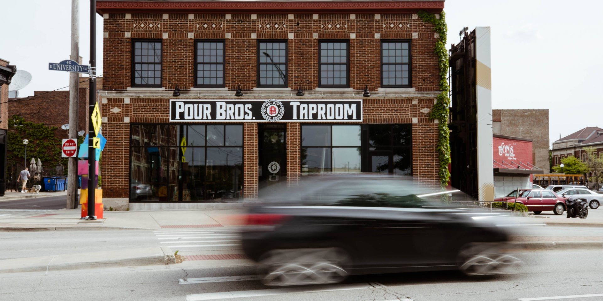 The exterior of Pour Bros. Craft Brewery for the Market Street Beer Fest in Champaign, Illinois. There is a brick building with Pour Bros on the outside above the door and a black car driving past is blurred. Photo by Anna Longworth-Singer.