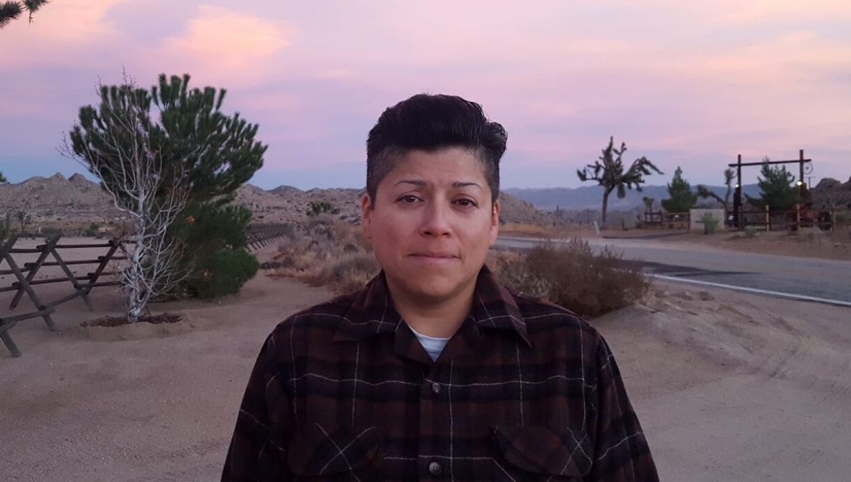 A person with dark hair and tan skin wears a dark plaid shirt with maroon stripes. They are standing in a desert with sand and a few trees. The sun has set and there is a pink sky behind them.