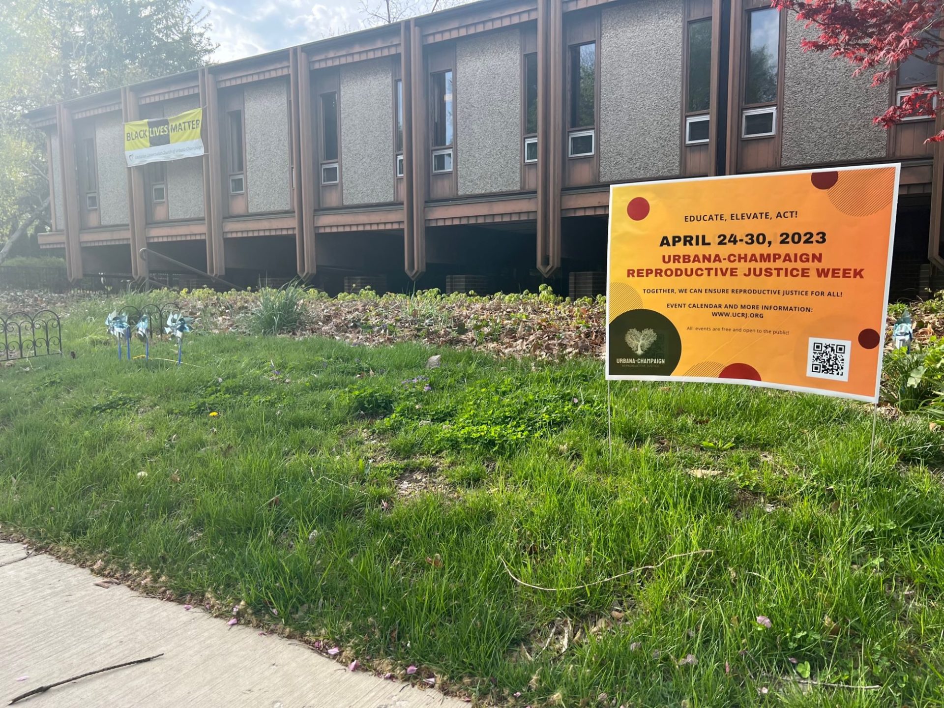 A golden yellow yard sign that says April 24-20 in black lettering, and Urbana-Champaign Reproductive Justice Week in red letters. It's in a patch of grass in front of a one story brown building with a row of large windows in front.