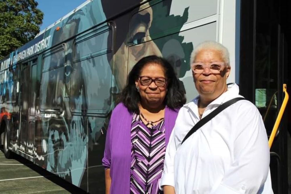 Two Black woman stand next to each other, alongside a city bus that is painted with an image of Frederick Douglass. One woman has shoulder length dark hair and glasses, and is wearing a purple cardigan and purple, white, and black striped shirt. The other has short white hair and glasses, and is wearing a white collared shirt with a black strap across the front.