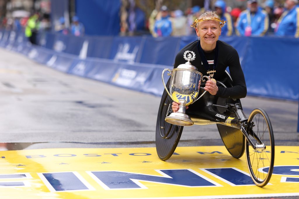 A white woman with blond hair, wearing a black track suit, is in a racing wheelchair. She is posed on a painted yellow finish line, holding a large gold trophy.