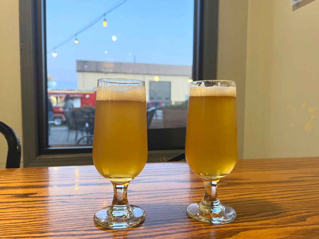 Two light colored beers in small glasses are on a wooden table inside Triptych Brewing. There is a red food truck named Marrakesh Grill outside. Photo by Alyssa Buckley.