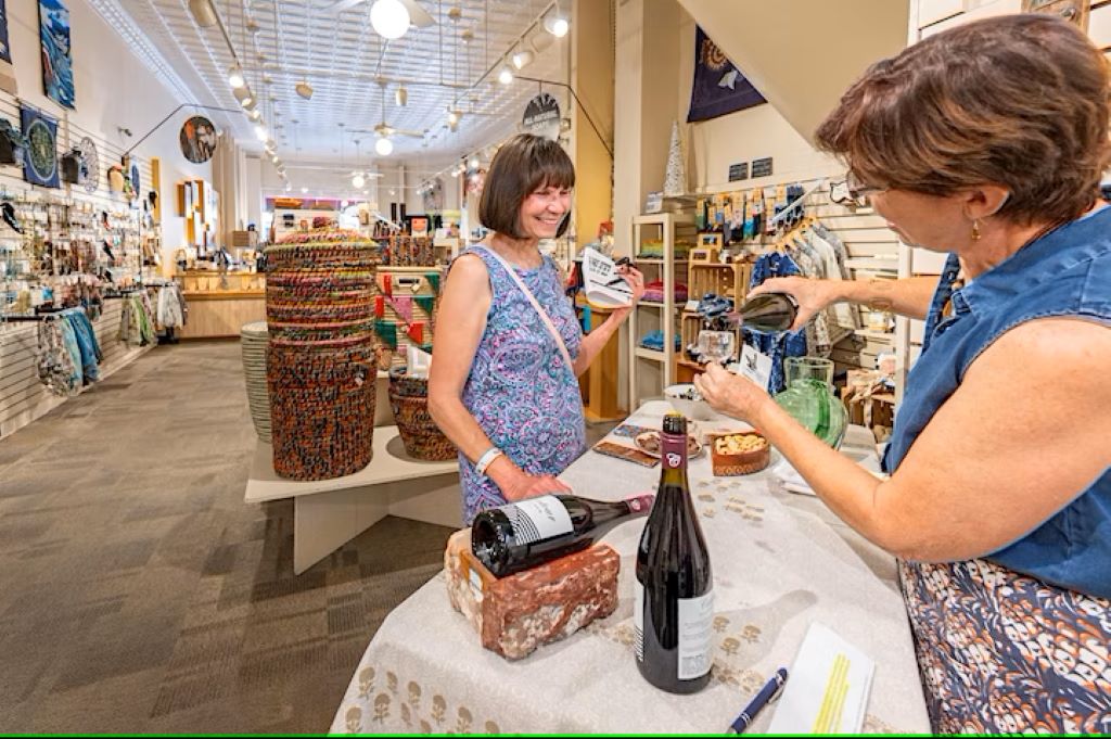 A white woman with short brown hair is standing behind a small table with a white tablecloth. She is pouring a class of wine for another white woman with short brown hair. They are in a retail store.