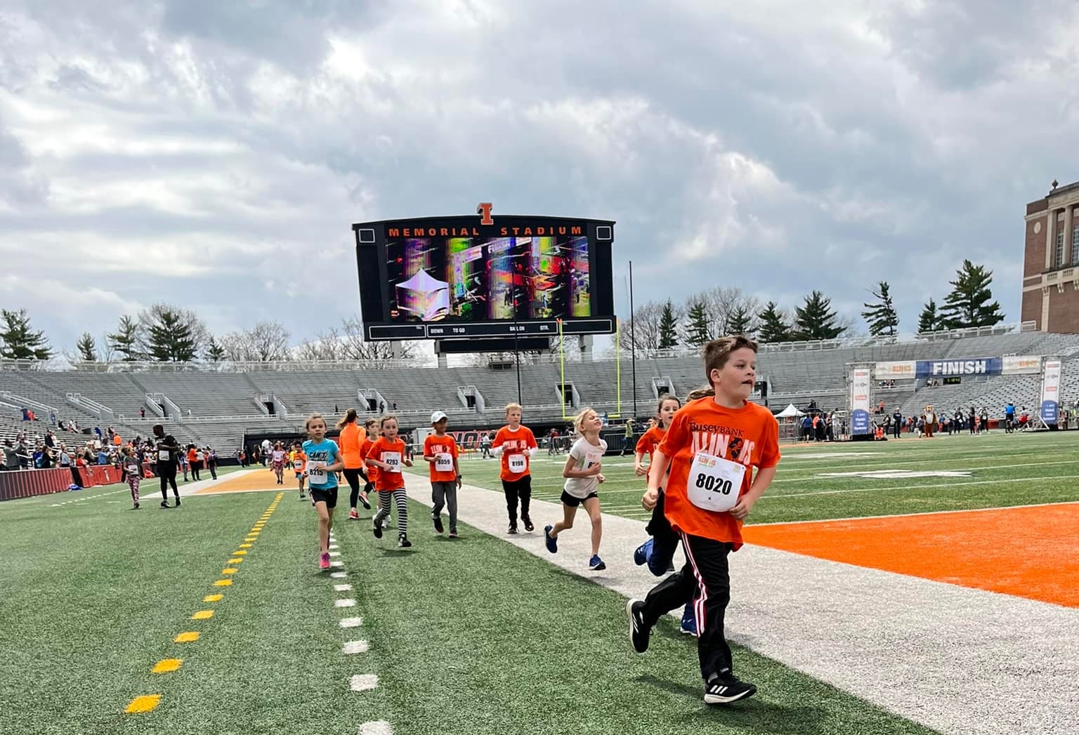 Several children in orange t-shirts are running along the sideline of a football field.