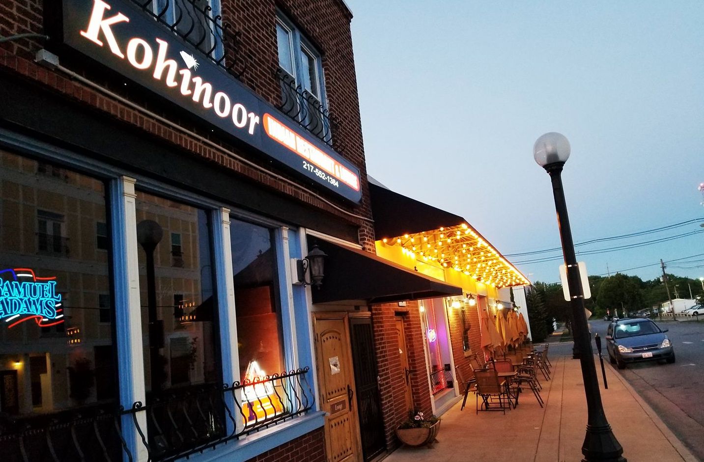 The outside of Kohinoor Indian restaurant at night. A large sign says Kohinoor above the door, next door there are white light strands hanging from the roof.