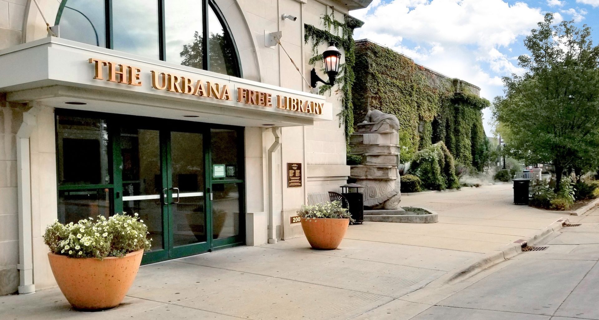 an outdoor shot of the urbana free library, a large white building with green doors, and a moss covered wall in the background