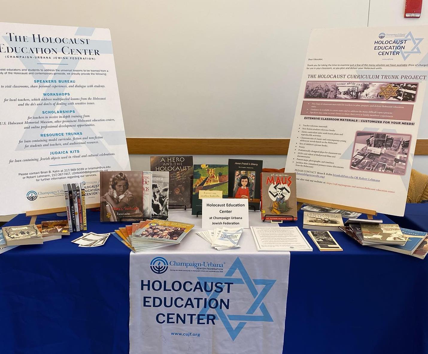 A blue display table. There is a sign hanging on the front saying "Holocaust Education Center." On the table are two large information posters, and various books including "Maus" and "Anne Frank's Diary," amongst others.