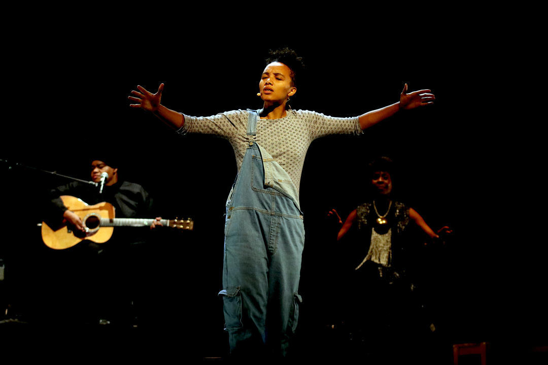 A Black woman stands on stage with her arms stretched out. She is wearing a grey long sleeve shirt and overalls, with one strap hanging down