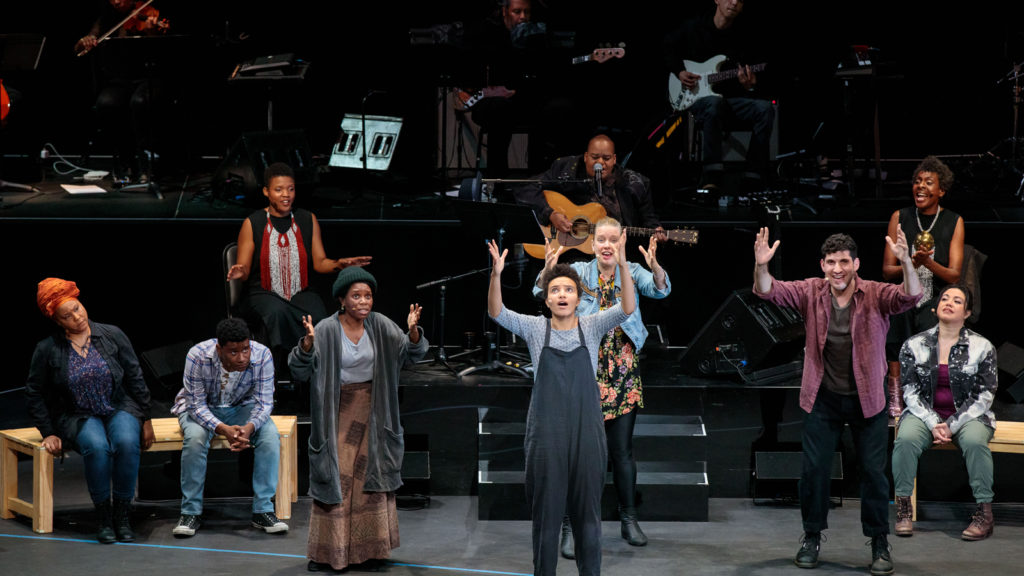 The cast of Parable of the Sower on stage; four cast members stand with their arms outstretched to the sky as they sing. Musicians are visible behind them