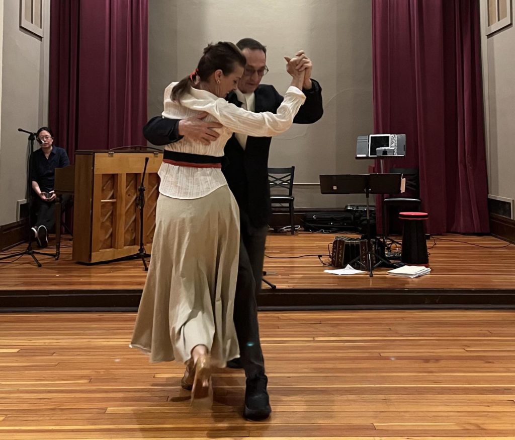 Joe Grohens dances tango with Carlota Bullard. Grohens wears a black suit and Carlota wears a long gold-ish skirt and white long sleeve top.