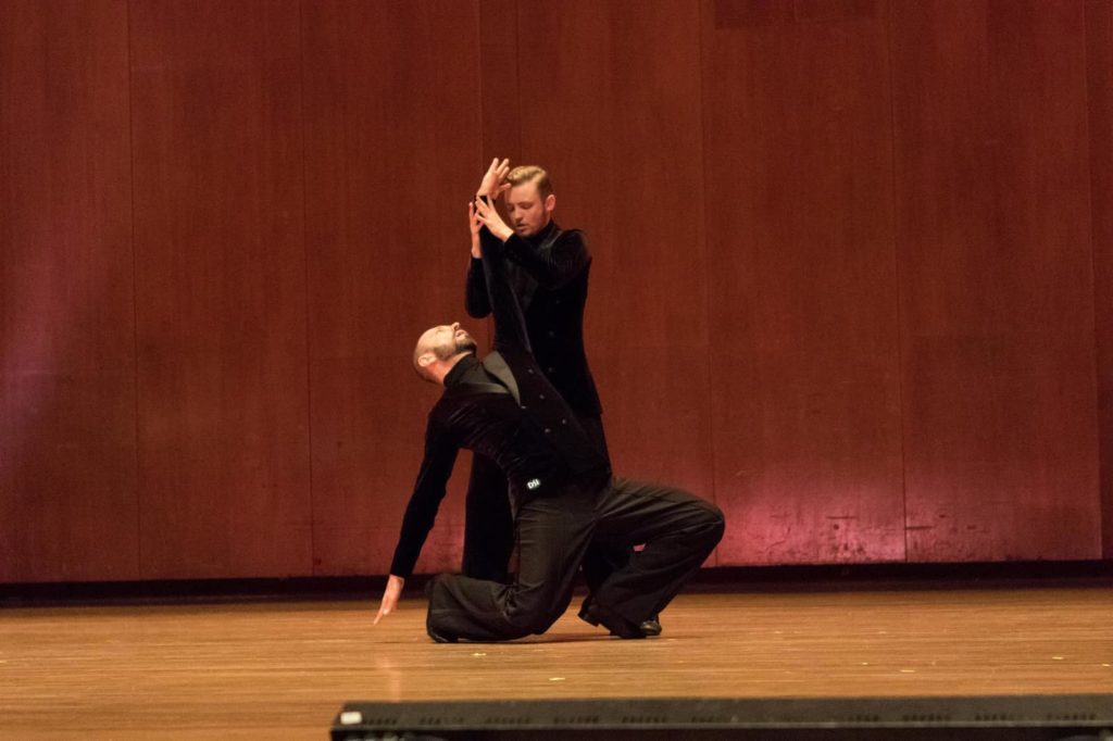 Tecza and Lindholm dance on stage wearing matching black pants and long sleeves. Tecza is on the floor with one hand touching the ground behind him and the other extended up, and Lindholm stands above with grasping the raised arm. 