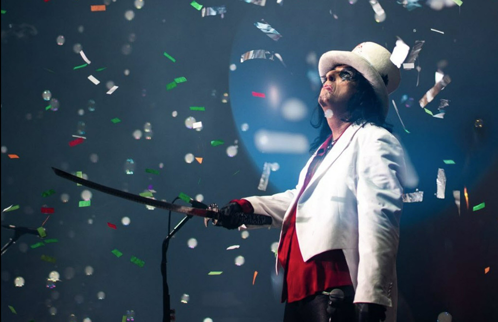Music artist Alice Cooper in a white and red suit, wearing a white top hat, looking to the sky while confetti rains down. He is holding a sword straight out.