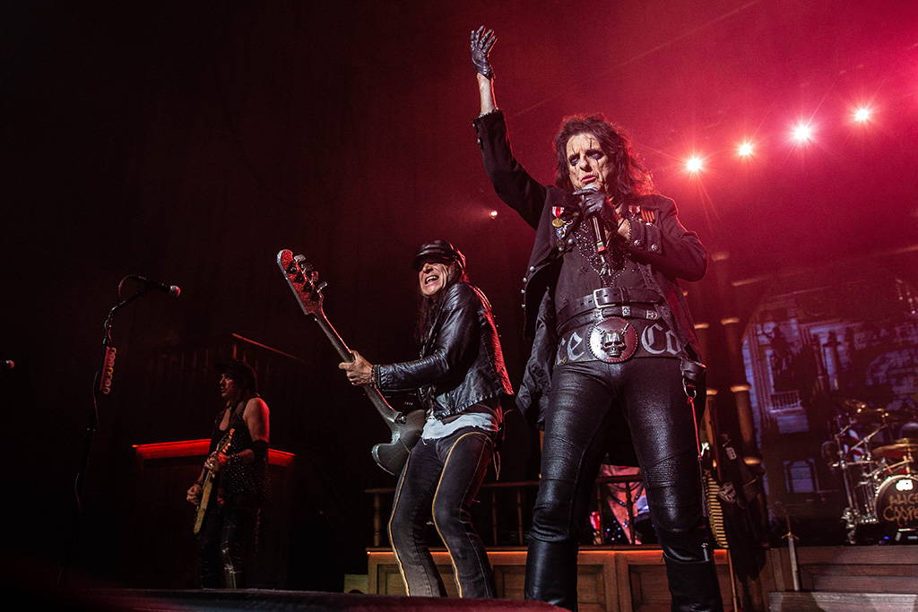 Alice Cooper performing onstage flanked by his bass player on his left. Off in the far left is another of Alice Cooper's guitarists.