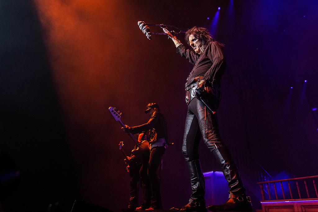 Alice Cooper and his band performing under a reddish light.