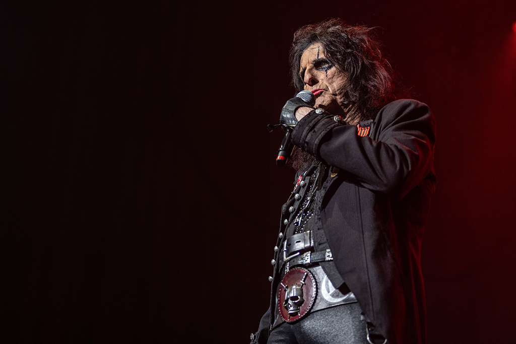 Alice Cooper, dressed in black, looking out over the crowd with the microphone to his mouth. He has a skull on his belt buckle.