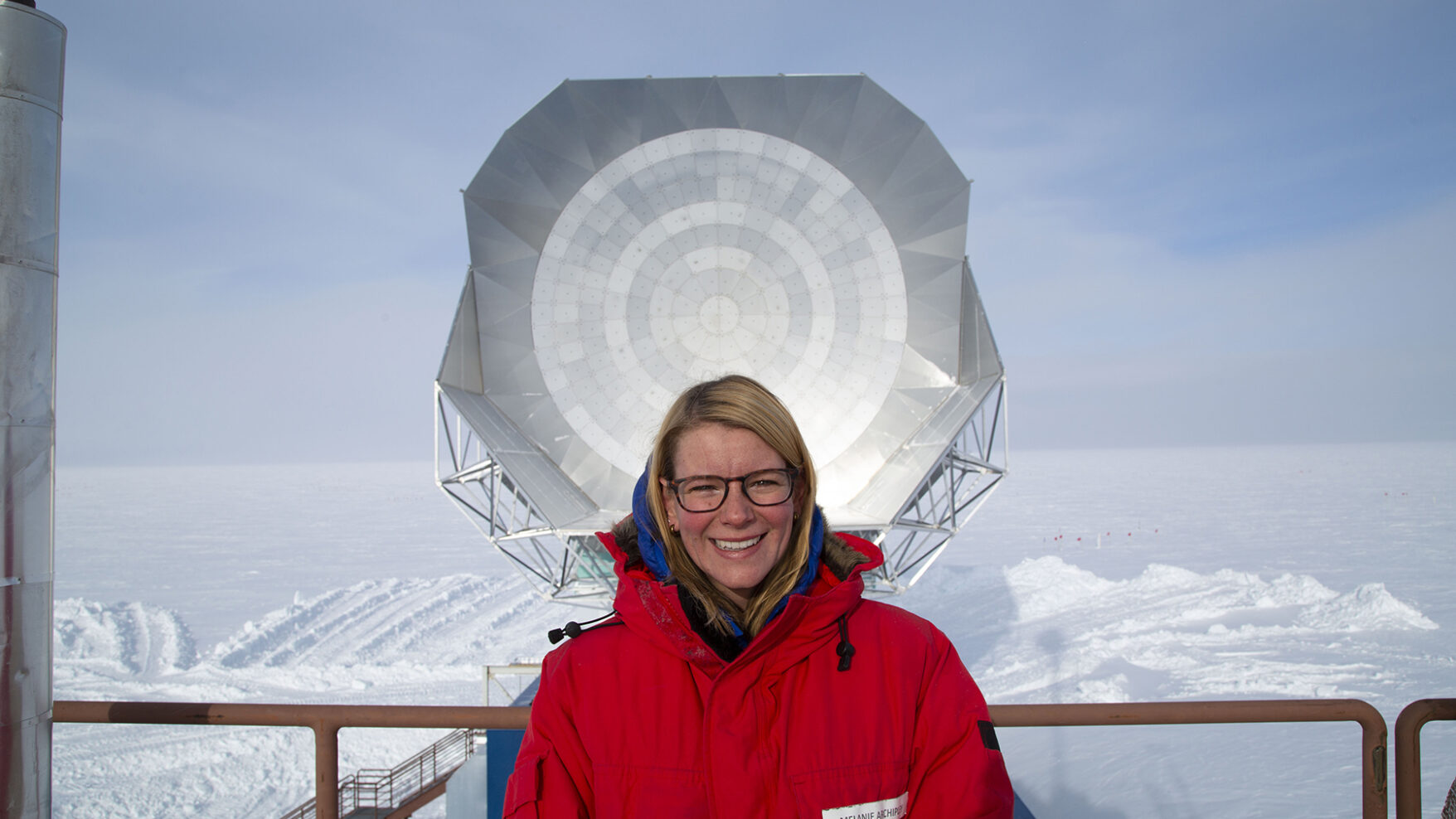 A white woman with blond hair, glasses, and a red coat is standing in front of a large white disc. In the background is a vast landscape of snow and ice, and blue sky.