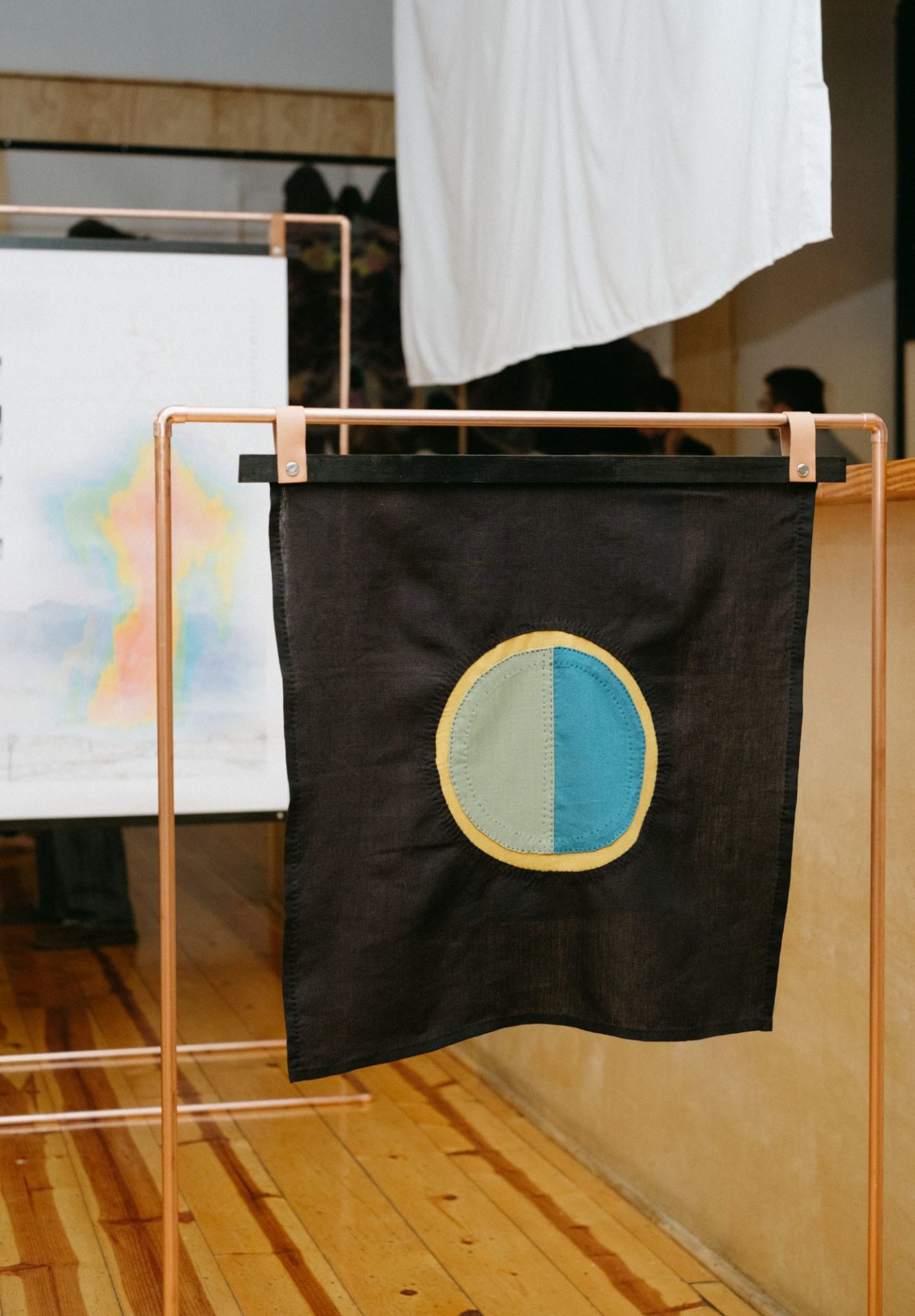 A circle that is half taupe and half teal with a gold border is stitched onto a rectangular black piece of fabric. The fabric is hanging on a frame made of copper tubing.