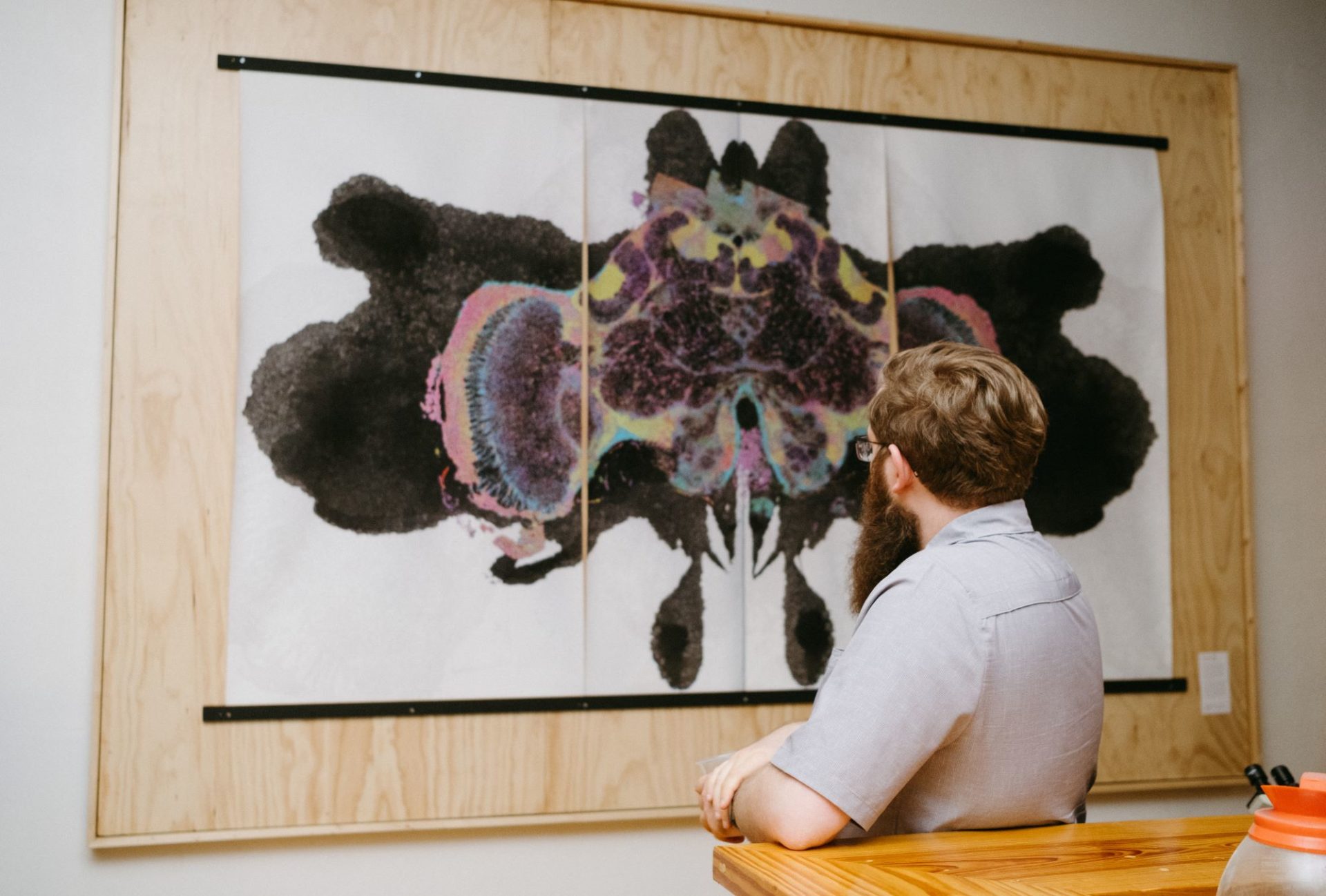 A large printed image, reminiscent of a Rorshach test, with a white backround. It is hanging on a wooden backdrop. A man with long beard and glasses is leaning against a wooden countertop and looking at the image.