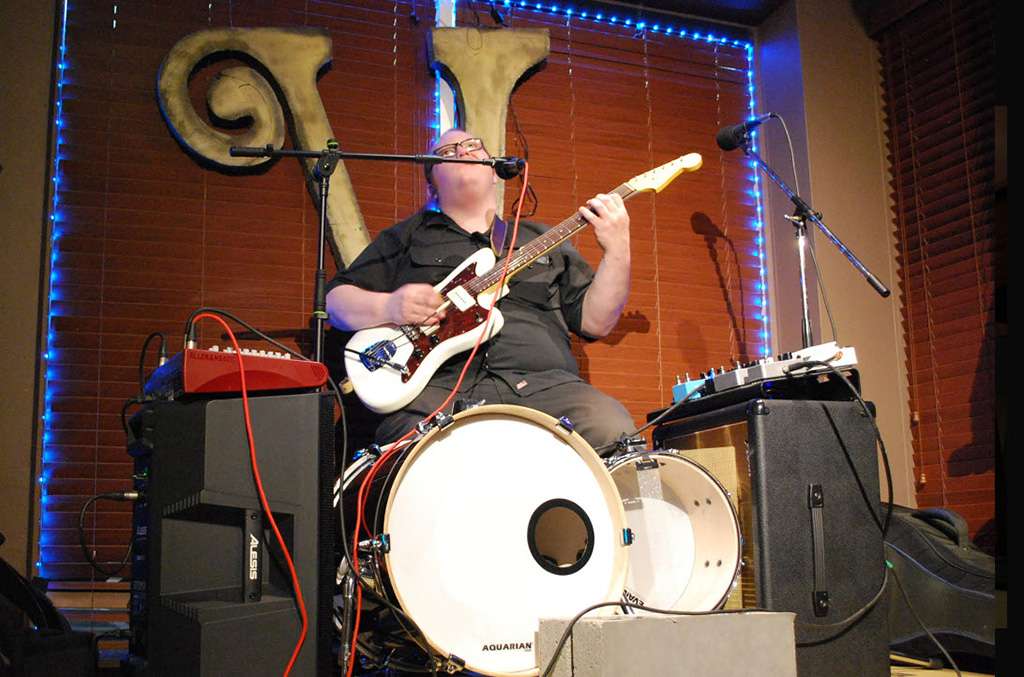 One man band Bobcat onstage, holding a guitar with kick drums below him, a pedal board on an amp to the right, and a P.A. system sitting on a monitor set on its side to the left. 