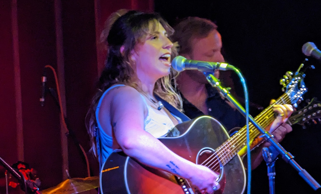 Singer Carrie Chandler of Carrie Sue and the Woodburners performing onstage. She is in a white sleeveless top and playing a guitar while singing into a mic. The band's mandolin player is in the background with a black shirt on, and playing his mandolin.