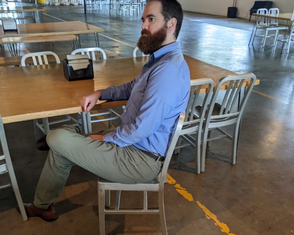 a white man with brown hair and a red beard sits comfortably on a metal chair. He is wearing a blue button up long sleeve shirt and khaki pants. He has one arm resting on the chair next to him.