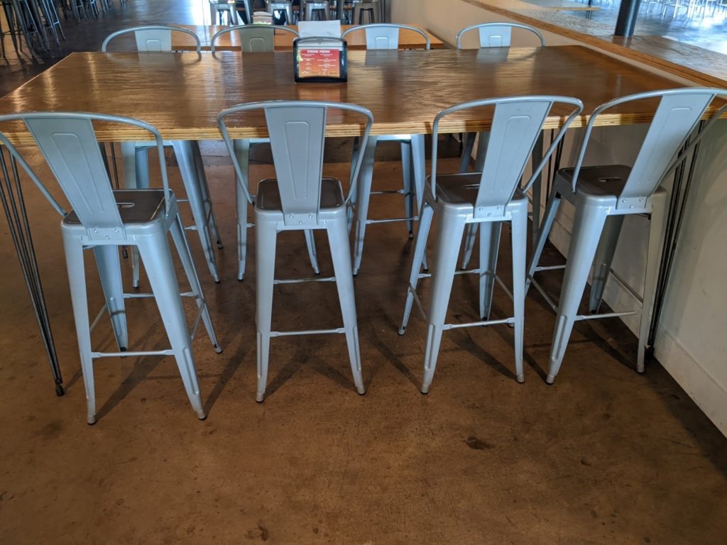 Four gray metal chairs sit pushed under a wooden table jutting out from a wall of windows. The floor is brown.