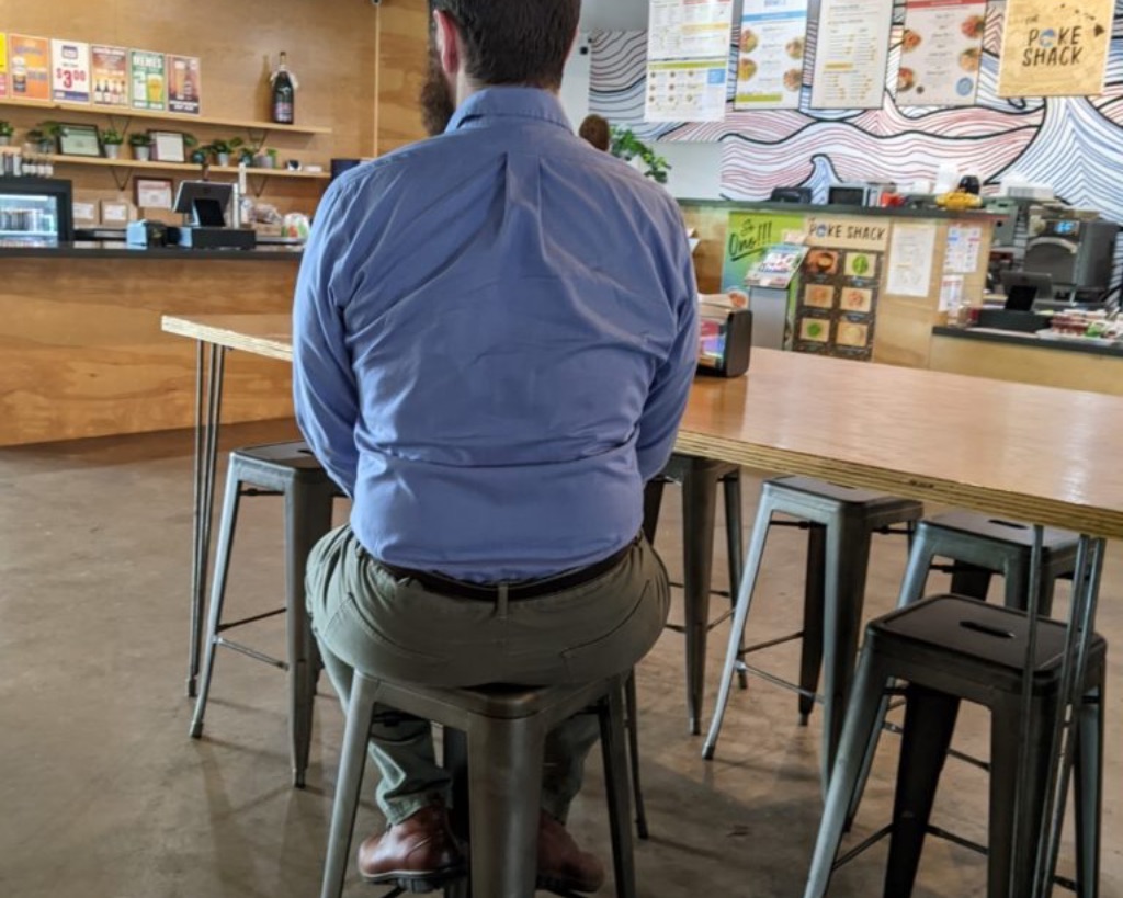 A picture of a white man with brown hair from behind. He sits snugly on a tall metal stool. He is wearing a blue button up long sleeve shirt and khaki pants. He looks uncomfortable.