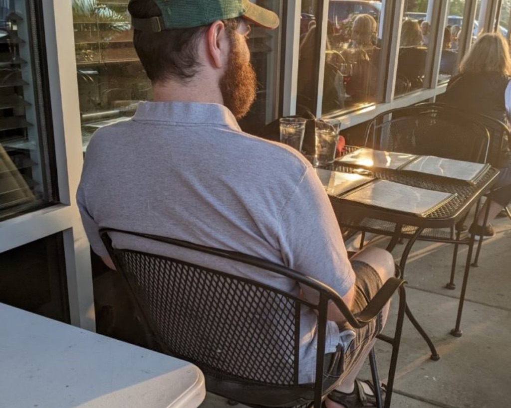 A side view of a white man with brown hair and a red beard sits comfortably in a black metal chair. He is wearing a gray polo shirt and a green baseball hat. The sun is shining on his face. 