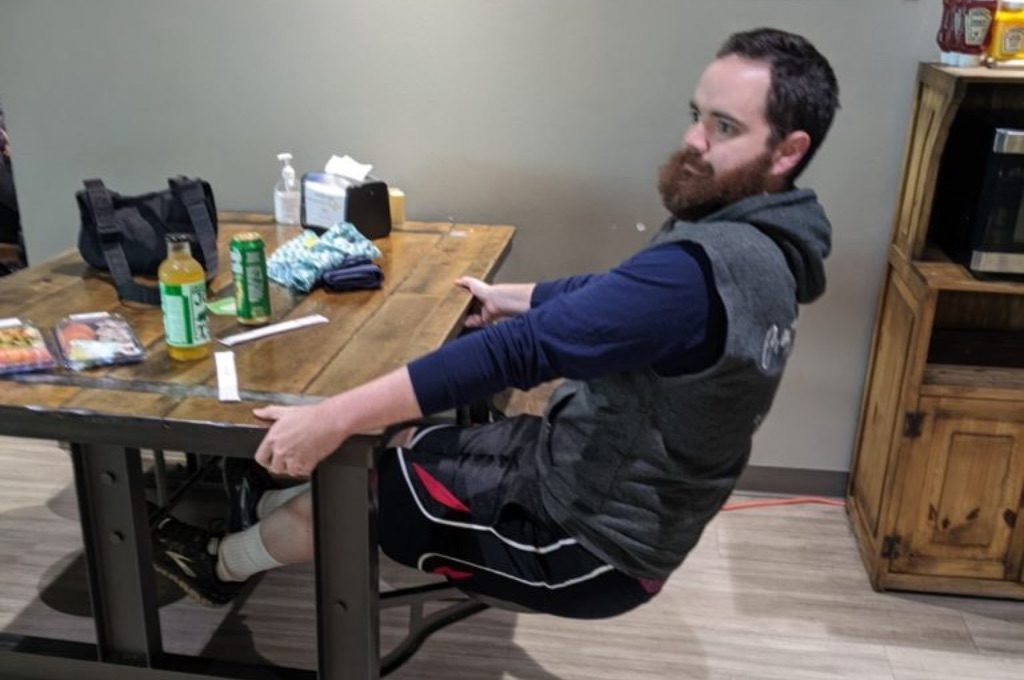a white man with brown hair and a red beard sits at a dark wooden table with a circular stool seat jutting out from each of the tables black metal legs. The wall is green. He is holding the table and wearing a blue and gray sweatshirt and dark blue shorts.