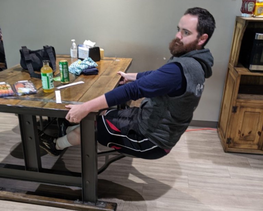 a white man with brown hair and a brown beard sits at a dark wooden table with a circular stool seat jutting out from each of the tables black metal legs. The wall is green. He is holding the table and wearing a blue and gray sweatshirt and dark blue shorts.